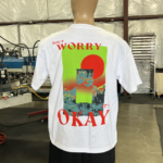 A mannequin wearing one of the 100 Garment-Dyed Heavyweight Shirts - Text-Based Artwork with Vector Designs - Maximum of 2 Ink Colors - Soft Hand Screen Printing, featuring vibrant red and green graphic text that reads "DON'T WORRY IT'S OKAY" in a workshop setting.